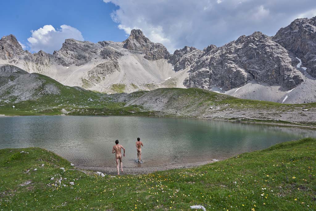 Das Bild eigt eine Szene zum Thema Klettern und Baden in Tirol. Zwei nackte Männer laufen in den Steinsee, im Hintergrund die Felswände.