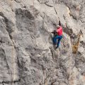 Zu sehen ist ein Bild zum Thema "Erstbeghung im Wetterstein". Ein Kletterer mit rotem Shirt klettert in einer grauen Felswand.