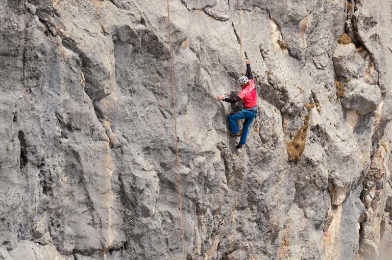 Zu sehen ist ein Bild zum Thema "Erstbeghung im Wetterstein". Ein Kletterer mit rotem Shirt klettert in einer grauen Felswand.
