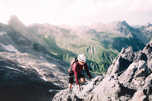 Eine Frau geht, gesichert durch ein Klettersteigset einen Klettersteig. Im Hintergrund sieht am die Tiroler Berge