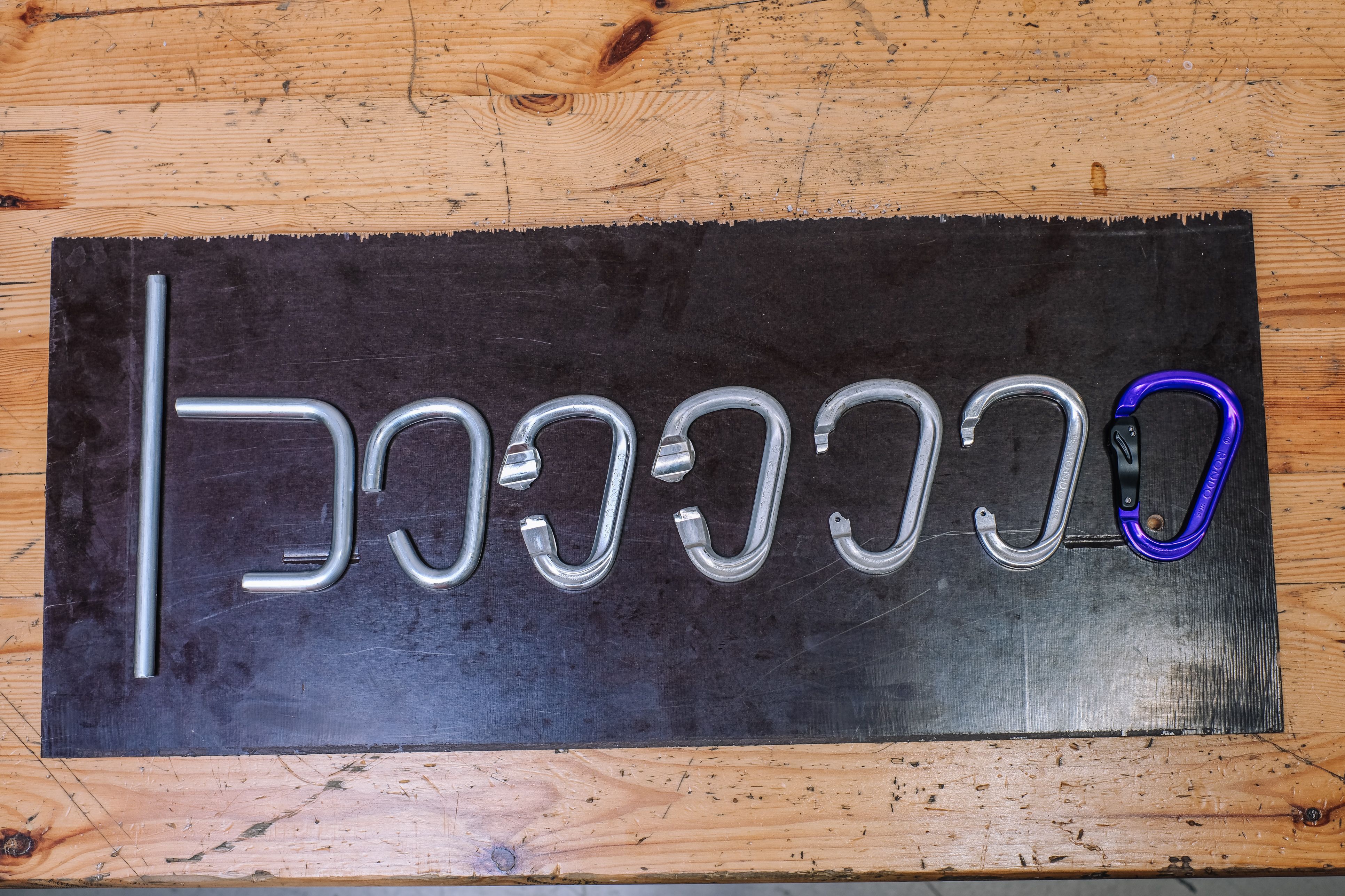 Aluminiumstab, mehrere Zwischenstufen der Fertigung und ein fertiger violetter Karabiner nebeneinander auf einer Holzplatte