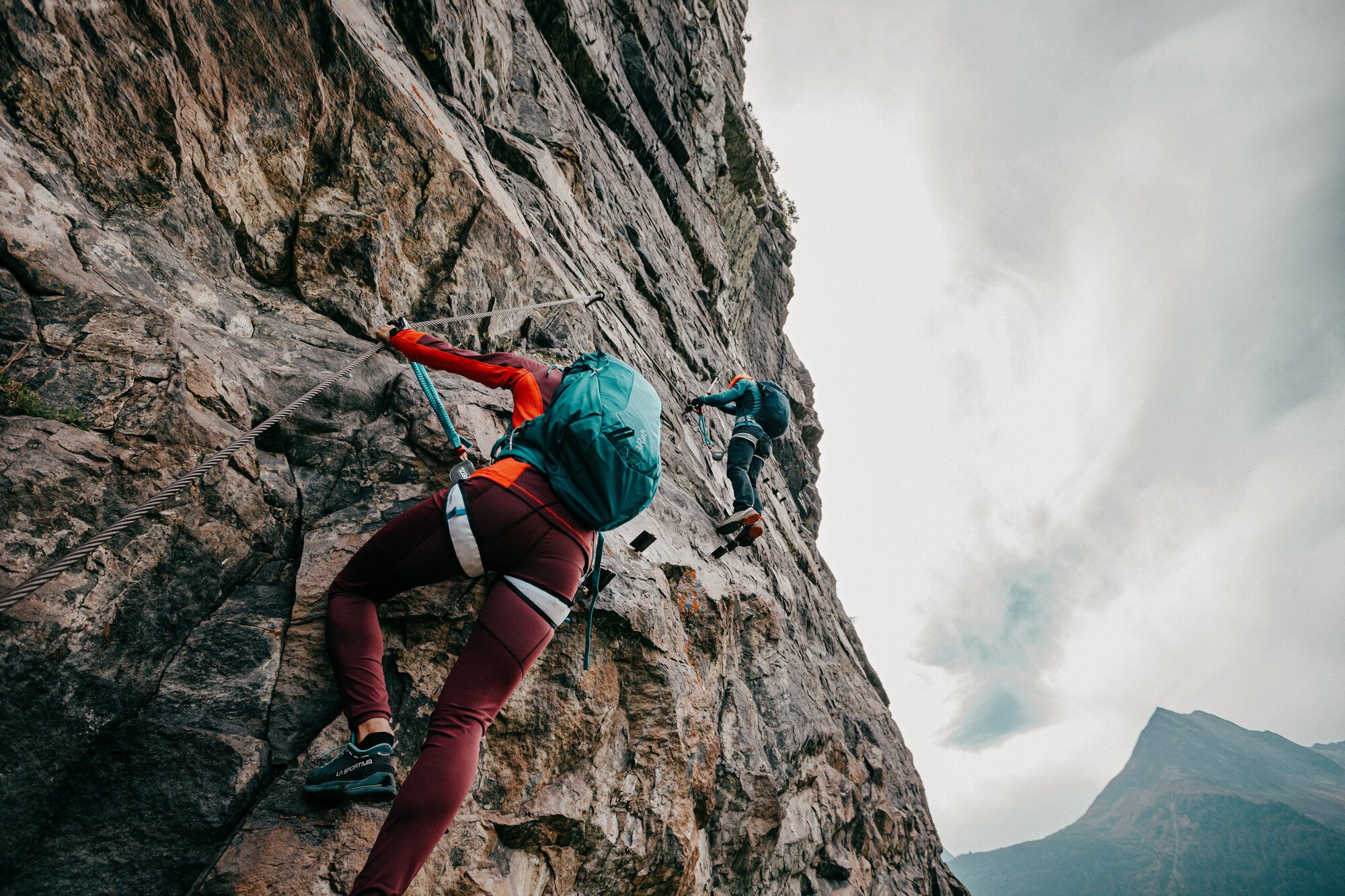 Eine Frau bewältigt gerade eine schwierige Passage am Klettersteig. Ein Mann schaut ihr von einer Stelle, die weiter oben liegt zu.