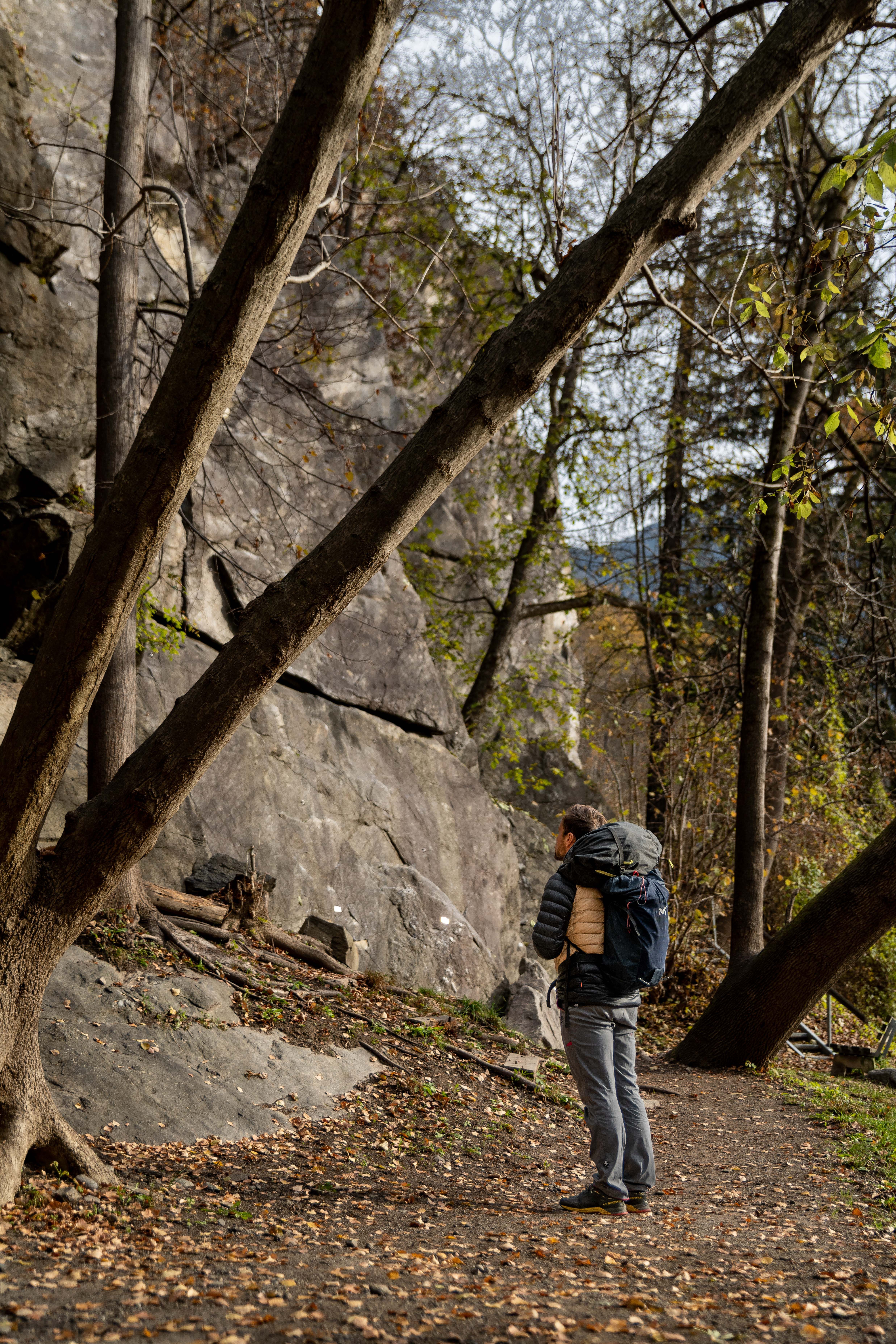 Person mit Rucksack am Wandfuß des Klettergartens Oetz zwischen Bäumen, Blick zur Wand