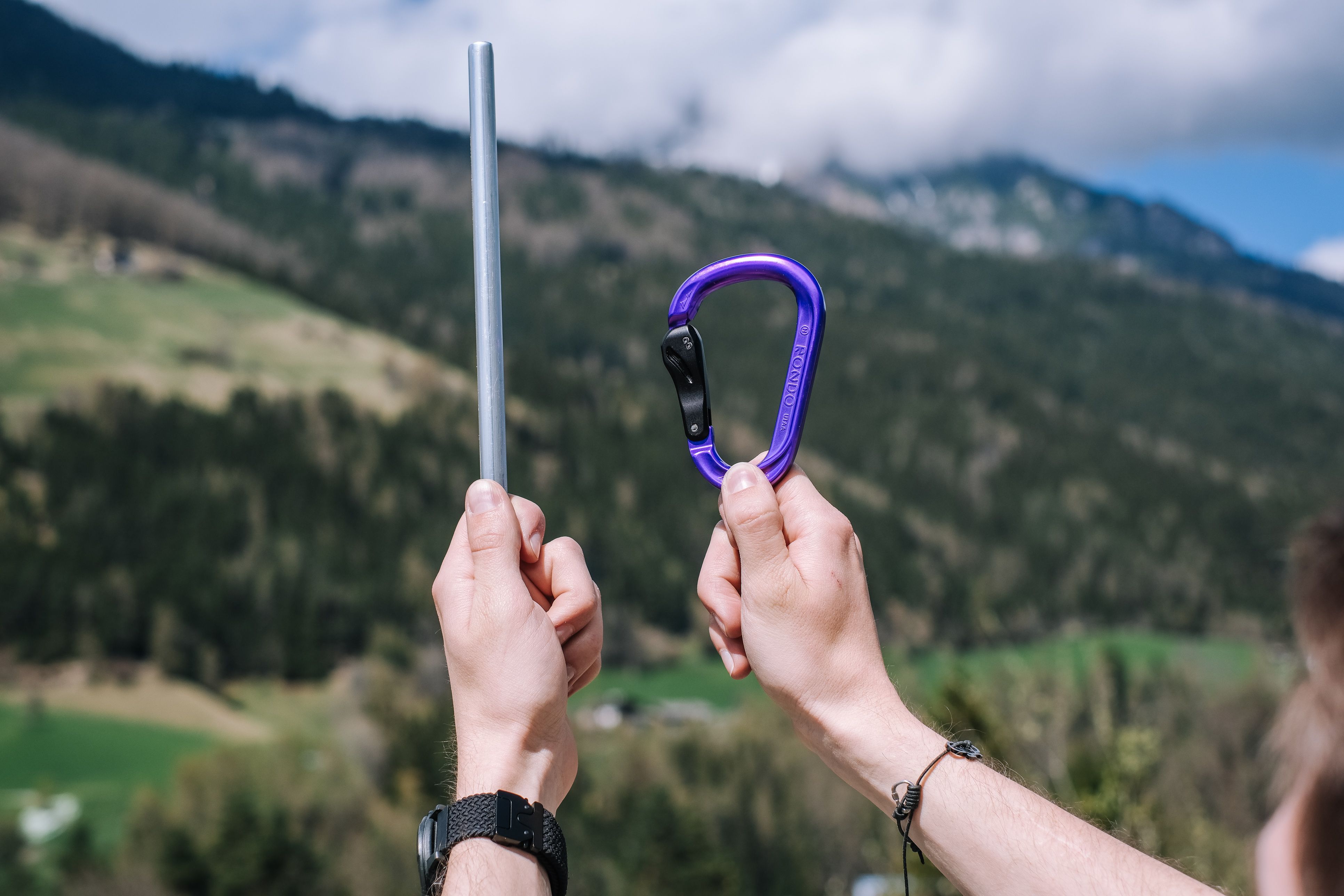 Zwei Hände halten vor Berglandschaft einen Aluminiumstab und einen fertigen violetten Karabiner