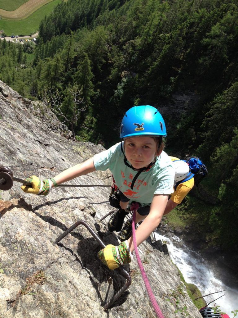 Kind mit Helm, Klettergurt und Klettersteigset an einer senkrechten Felswand